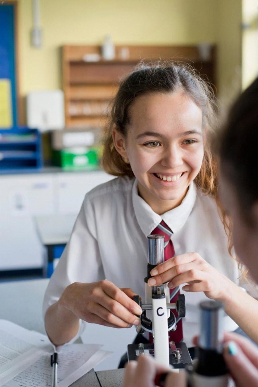Smiling primary school girl in uniform using a microscope during a practical science lesson